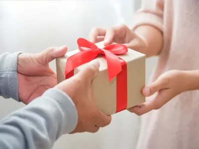 Eine schöne Geschenkbox wird von Händen in andere Hände überreicht. Eine rote Schleife an der Box. Ein Geschenk der besonderen Art